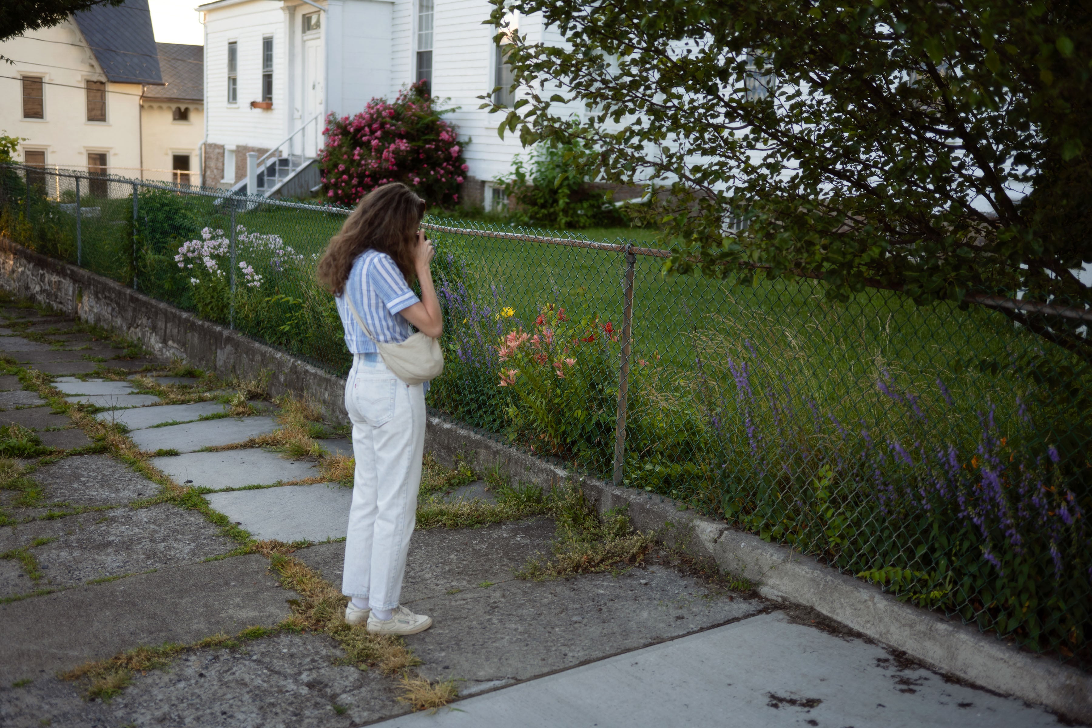 A young woman standing outside a house photographing flowers